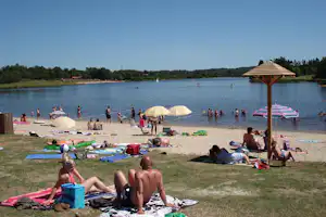 Badebereich am See mit Liegen, Sonnenschirmen und mehreren Personen im Wasser, im Hintergrund Wald und Ufer.