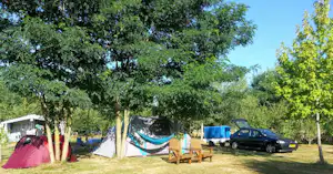 Campingzelt mit Hängematte, zwei Stühlen und Tisch auf dem Stellplatz, daneben geparkter Pkw und Wohnwagen im Grünen.