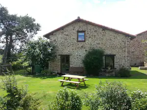 Steinhaus mit Terrasse und Picknicktisch im Campinggarten, umgeben von Rasen, Büschen und Bäumen.
