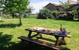 Garten am Campingplatz mit Holztisch und Grillgut, daneben Flasche und Glas, im Hintergrund Bäume und ein Haus.