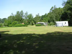 Campingwiese mit Wohnwagen und Vorzelten, im Hintergrund Bäume und ein kleines Gebäude.