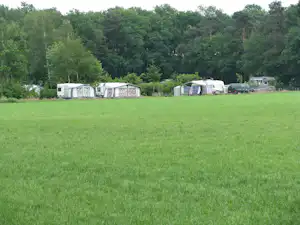 Wohnwagen und Zelte auf einem grünen Campingplatz, im Hintergrund Bäume und ein Parkplatz mit Fahrzeugen.