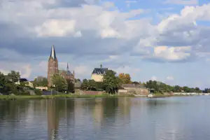Kirchengebäude und Schloss am Flussufer, mit Bäumen und Häusern im Hintergrund, aufgenommen über dem ruhigen Wasser.