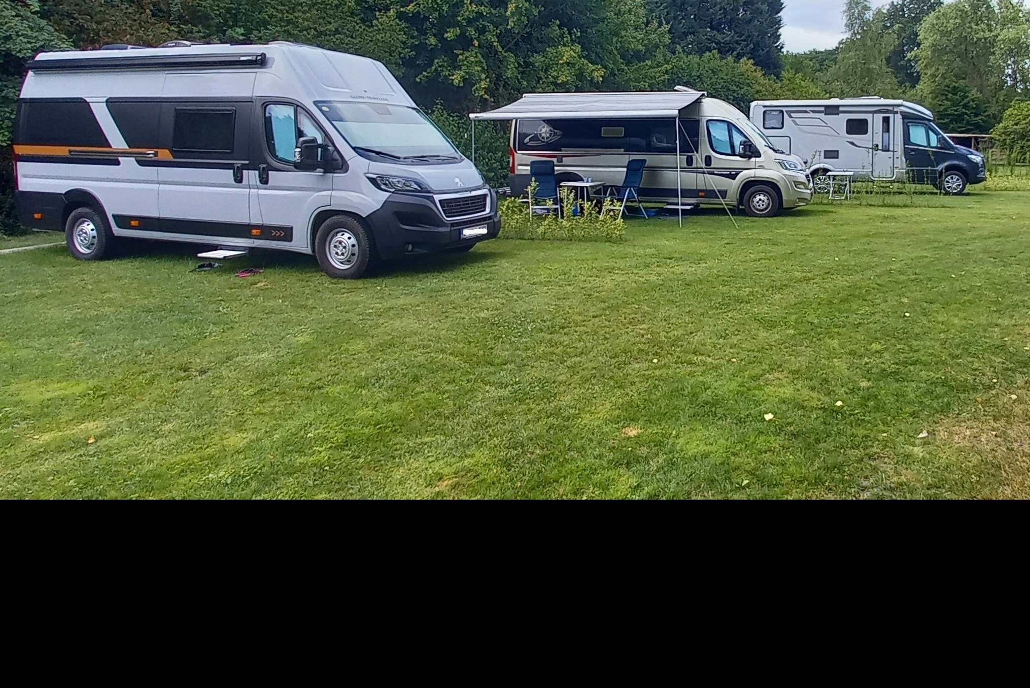 Wohnmobile auf einem grünen Campingplatz, mit Vorzelten und Tischen, im Hintergrund Bäume und ein weiterer Stellplatz.