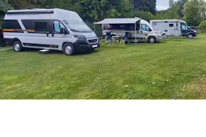 Wohnmobile auf einem grünen Campingplatz, mit Vorzelten und Tischen, im Hintergrund Bäume und ein weiterer Stellplatz.