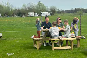 Familie sitzt an einer Picknickbank am Campingplatz, im Hintergrund Wohnwagen und Wohnmobile auf einer grünen Wiese.