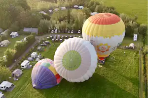 Zwei Heißluftballons über einem Campingplatz mit Wohnmobilen, Wohnwagen, Rasenflächen und Wegen zwischen Bäumen.