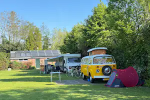Gelber VW-Bus und Wohnmobil auf dem Campingplatz, Zelt im Vordergrund, Solarmodule am Gebäude und Bäume im Hintergrund.