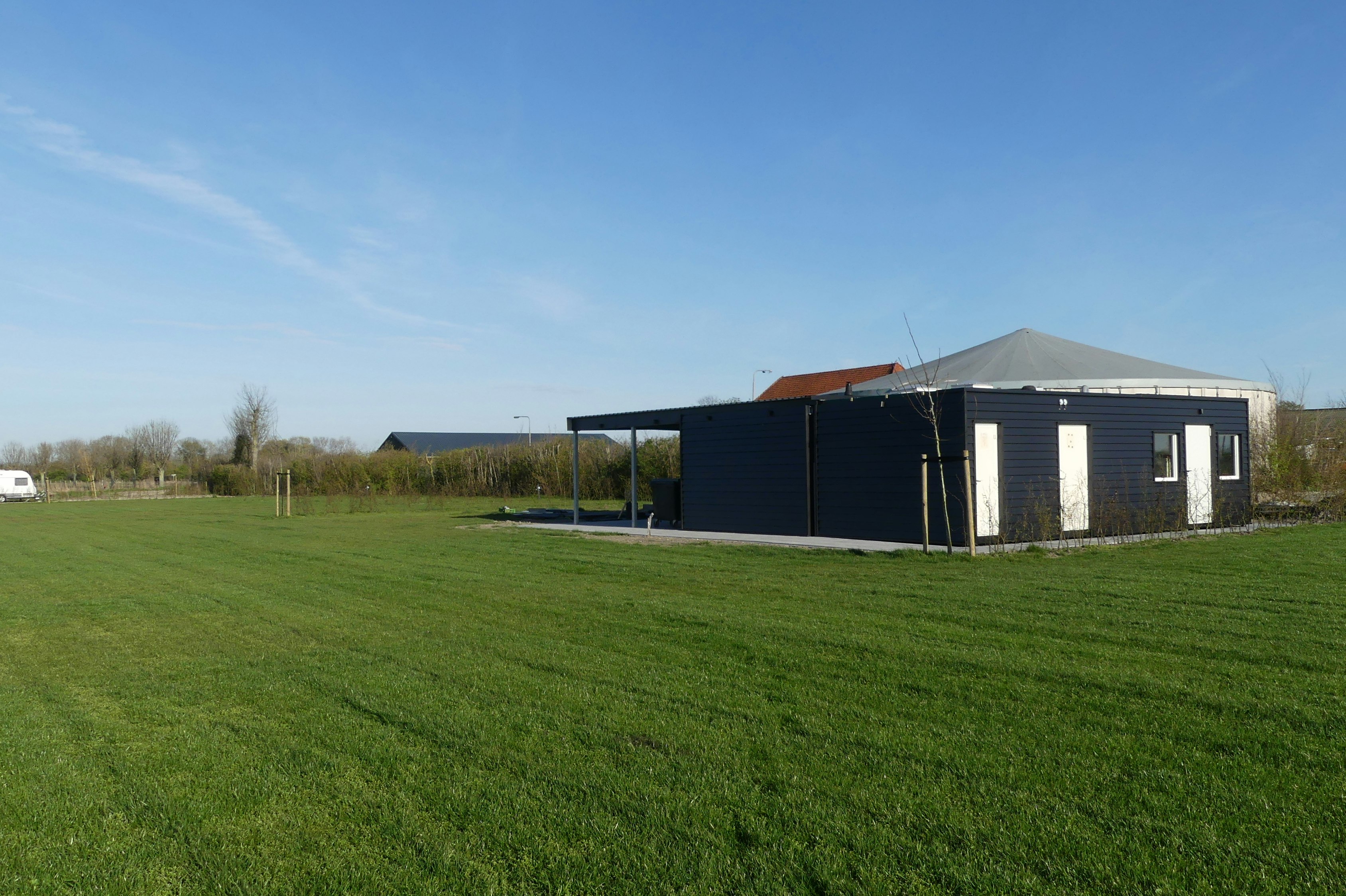 Campinghaus mit Carport und großen Fenstern, davor Rasenfläche und Hecke, im Hintergrund weitere Gebäude und Bäume.