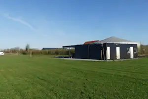 Campinghaus mit Carport und großen Fenstern, davor Rasenfläche und Hecke, im Hintergrund weitere Gebäude und Bäume.