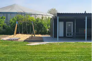 Camping-Unterkunft mit Terrasse, Holztisch und Schiebetür, daneben ein Spielbereich mit Schaukel und Zeltplane.