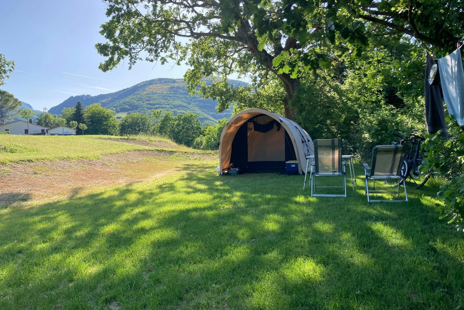 Zelt auf einem grünen Campingplatz mit Liegestühlen und Tisch, umgeben von Bäumen und Blick auf Hügel im Hintergrund.