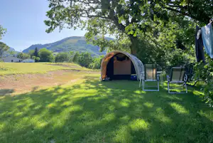 Zelt auf einem grünen Campingplatz mit Liegestühlen und Tisch, umgeben von Bäumen und Blick auf Hügel im Hintergrund.