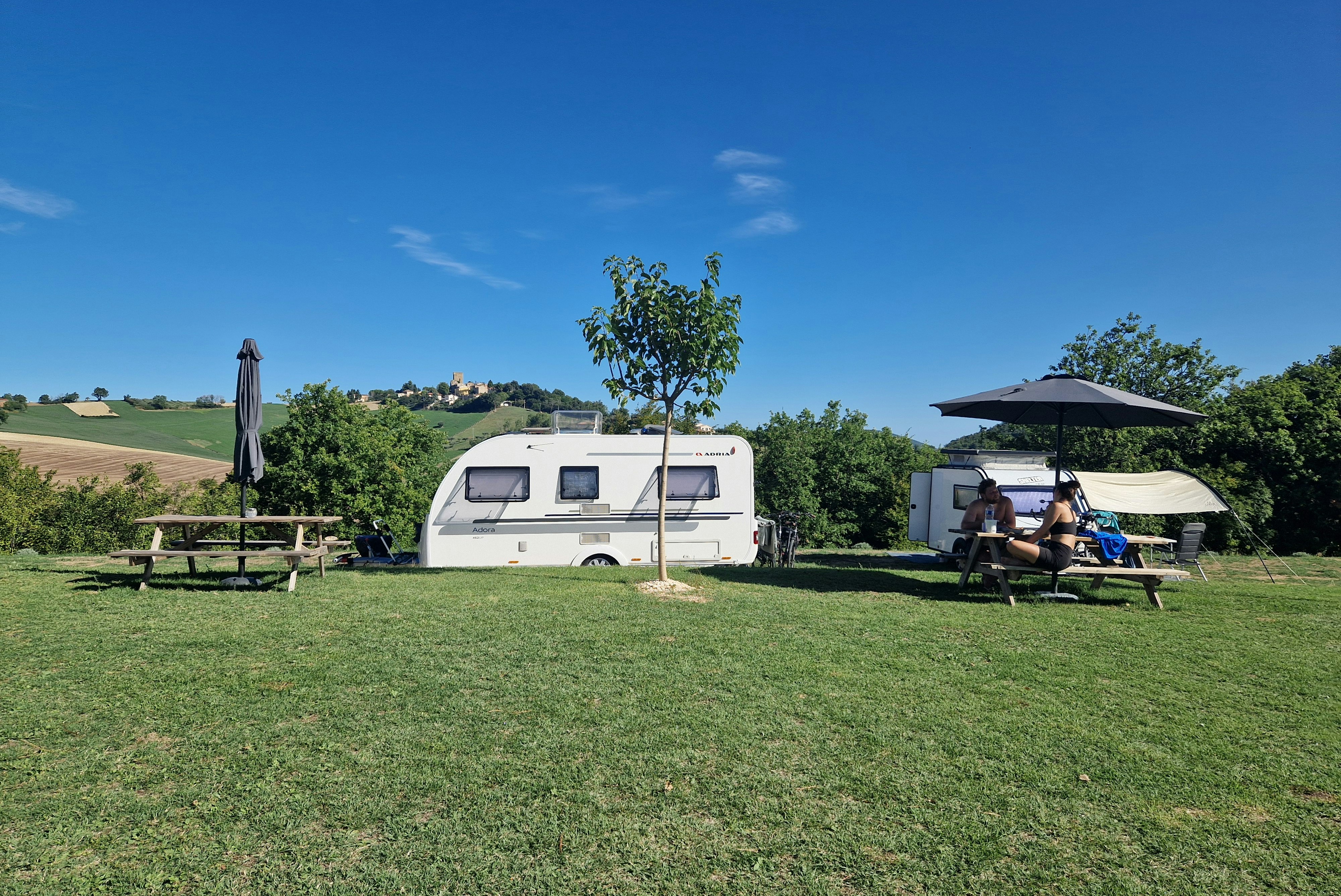Wohnwagen auf einer Campingwiese, daneben Sonnenschirm und Sitzbereich unter einer Markise, im Hintergrund Bäume und Hügel.