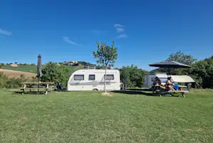 Wohnwagen auf einer Campingwiese, daneben Sonnenschirm und Sitzbereich unter einer Markise, im Hintergrund Bäume und Hügel.