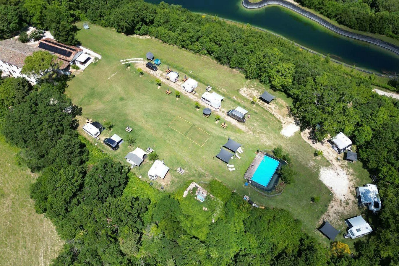 Luftaufnahme eines Campingplatzes mit mehreren Stellplätzen, Wohnwagen und Zelten, Wiesen, Wegen und einem blauen Pool am Rand des Sees.
