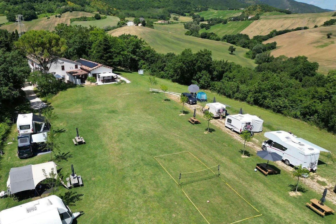 Luftaufnahme eines Campingplatzes mit Wohnmobilen, Zelten und Rasenflächen, umgeben von Bäumen und grünen Hügeln.