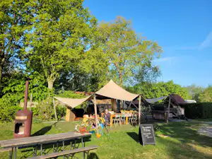 Zelte mit Sonnensegeln und Gartenmöbeln auf einer Wiese, im Hintergrund Bäume und ein Holzgebäude.