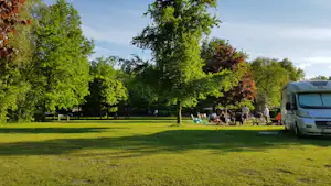 Große Campingwiese mit Wohnmobil am Rand, Bäume und Sitzbereich mit Stühlen im Hintergrund.