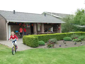 Campinghaus mit Terrasse, Hecken und Blumenbeeten; ein Kind fährt mit dem Fahrrad auf dem Rasen am Eingang vorbei.