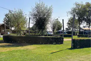 Grüner Rasen und Hecken im Campingbereich, dahinter Zäune und Bäume, im Hintergrund ein Wohnwagen und ein Weg.