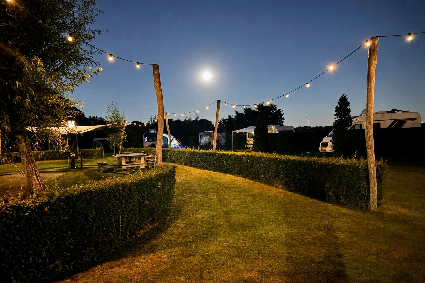 Gepflegter Campingplatz am Abend mit beleuchteten Wäscheleinen, Rasenwegen, Hecken und Sitzbänken zwischen Bäumen.