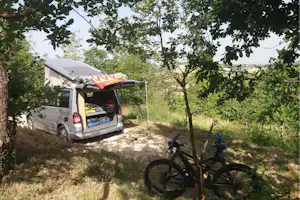 Wohnmobil mit geöffnetem Heck und Gepäck auf dem Stellplatz, im Grünen zwischen Bäumen und mit Fahrrad am Weg.