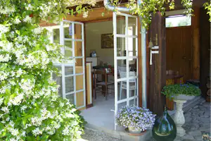 Terrasse am Campingplatz mit Holztisch und Stühlen, Glasfenster und Eingangstür, umgeben von Bäumen und Blumen.