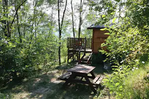 Holzhütte mit Terrasse und Sitzgruppe im Wald, umgeben von Bäumen und einem gepflasterten Weg.