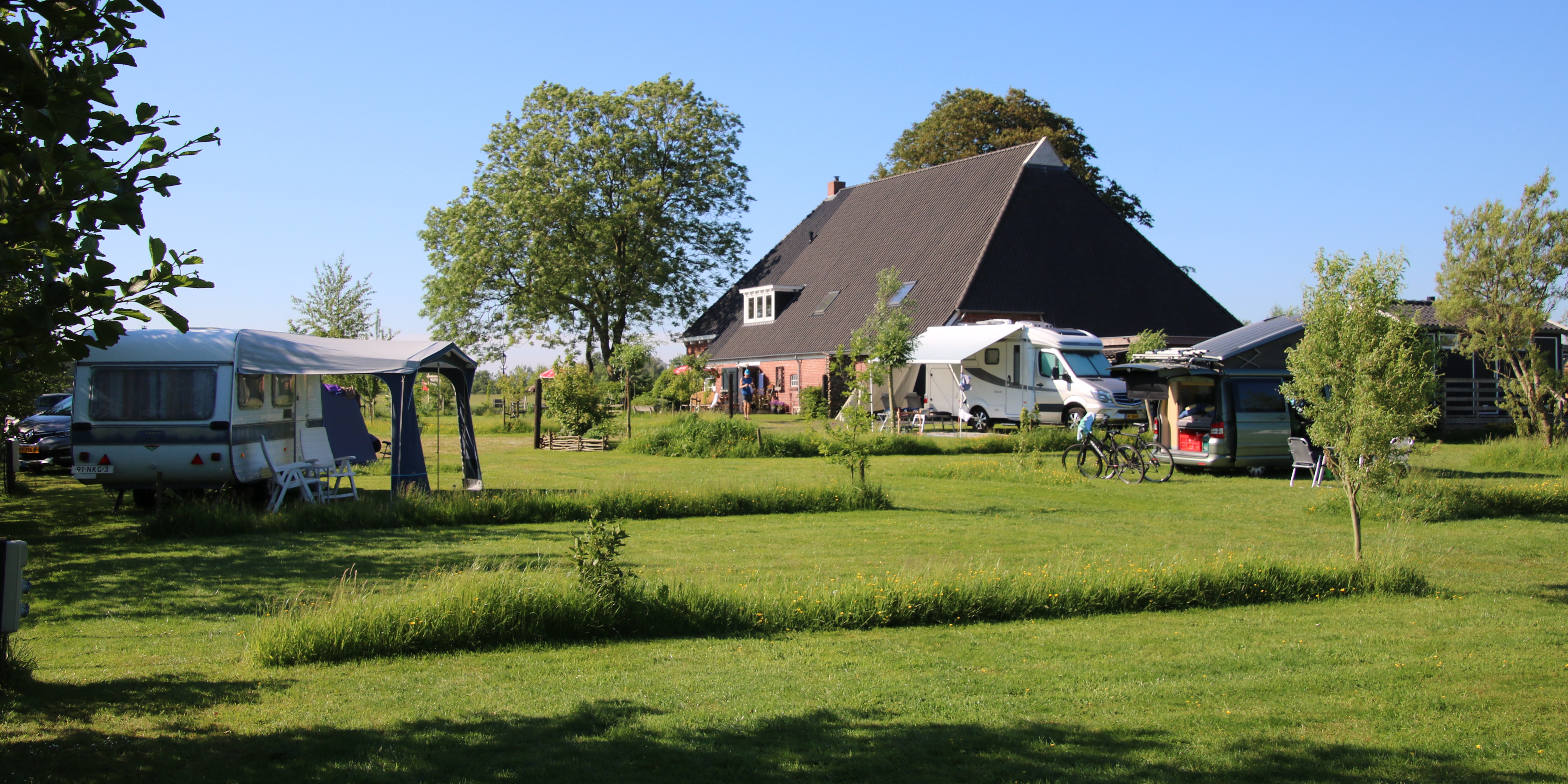 Campingplatz im Grünen mit Wohnwagen und Wohnmobilen auf dem Stellplatz, daneben ein Haus und Bäume im Hintergrund.