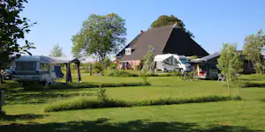 Campingplatz im Grünen mit Wohnwagen und Wohnmobilen auf dem Stellplatz, daneben ein Haus und Bäume im Hintergrund.