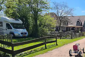 Wohnmobil am Campingplatz neben einem Holzzaun, im Hintergrund ein Haus mit Terrasse und Bäumen im grünen Garten.