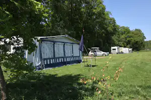 Campingzelt auf einer grünen Wiese, daneben ein Auto und weitere Wohnwagen unter hohen Bäumen.