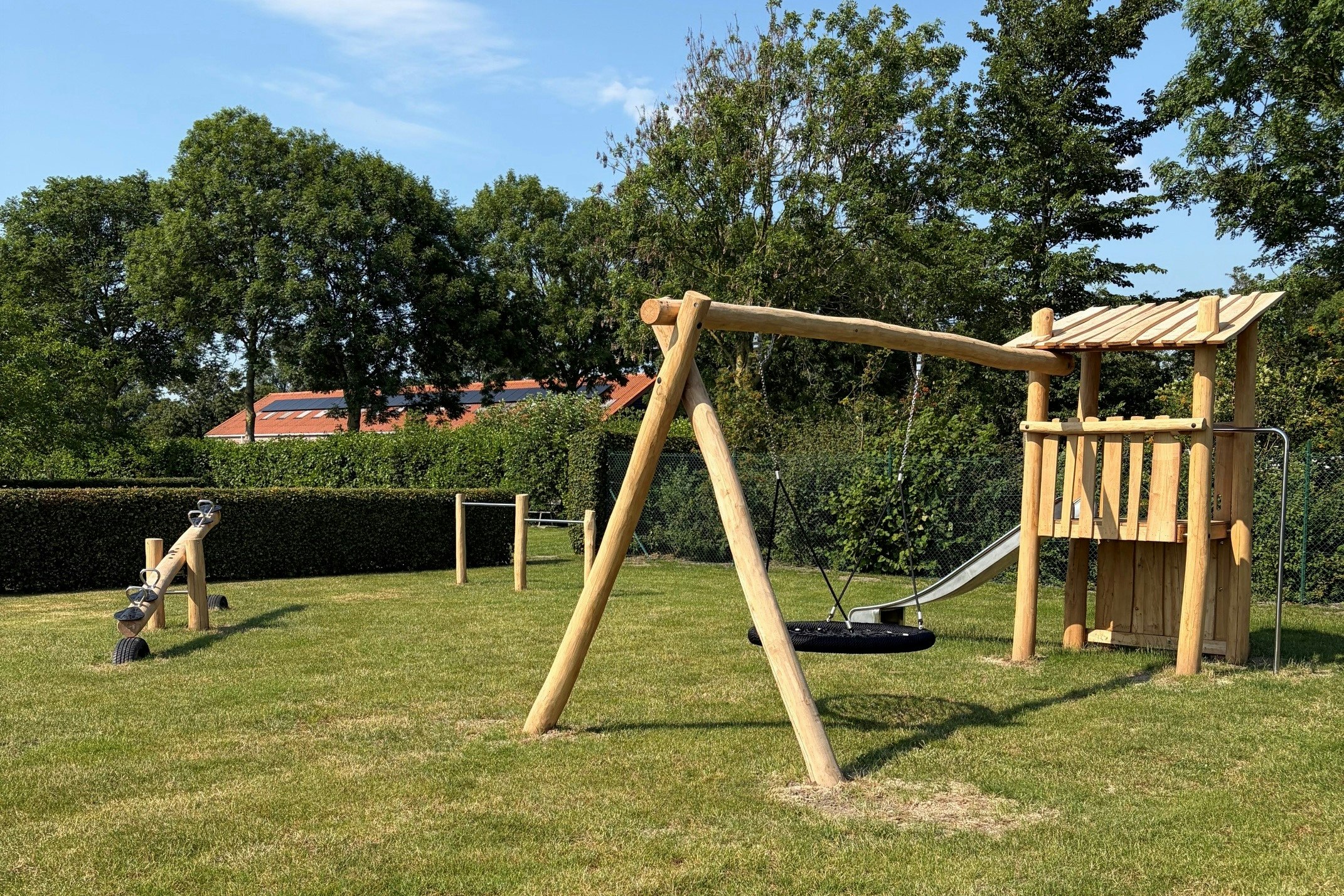 Spielplatz im Campinggarten: Holzschaukel und Klettergerüst auf Rasen, Hecken und Bäume im Hintergrund.
