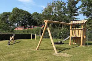 Spielplatz im Campinggarten: Holzschaukel und Klettergerüst auf Rasen, Hecken und Bäume im Hintergrund.