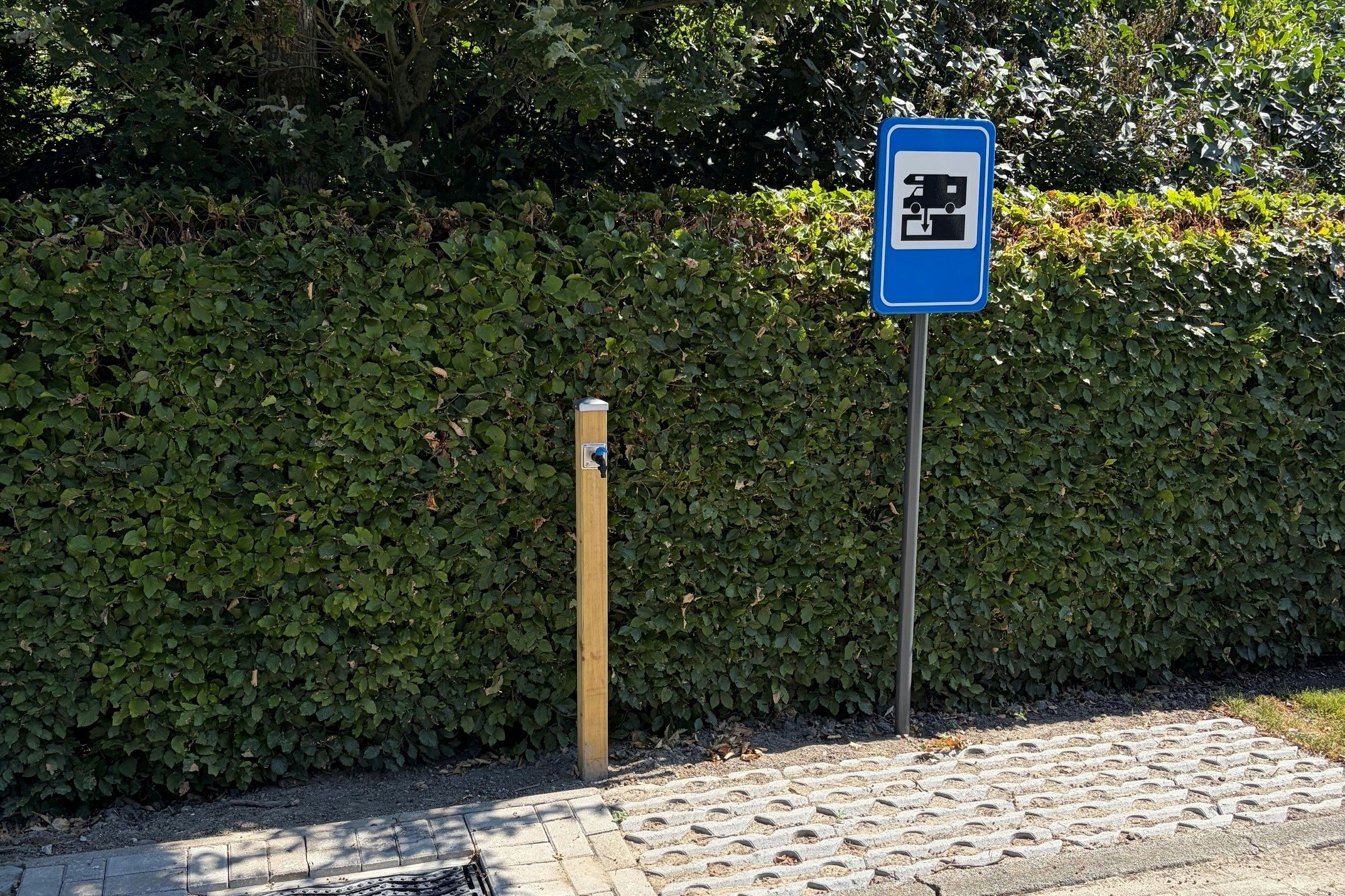 Blaues Camping-Schild an einem gepflasterten Weg, daneben ein Pfosten, im Hintergrund hohe Hecke und Bäume.