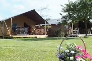 Zeltunterkunft mit Holzterrasse, Stühlen und Sonnenschirmen auf dem Campingplatz, im Vordergrund Blumen im Korb.
