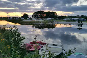 Ruderboot am Ufer, ruhiger See mit Spiegelung, im Hintergrund Zelte und Wohnwagen auf dem Campingplatz.