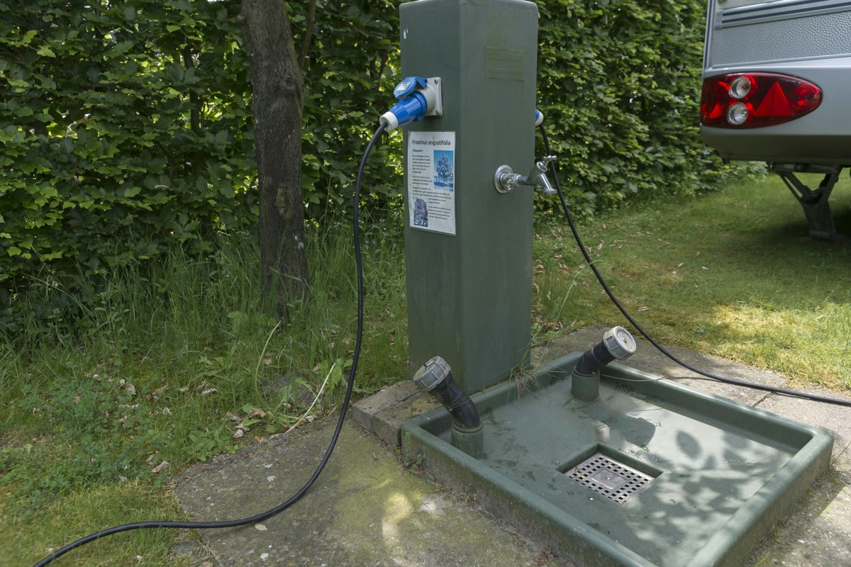 Wasseranschluss am Campingplatz mit Zapfhahn und Schlauch, daneben ein Spülbecken auf Beton, im Grünen mit Hecke und Rasen.