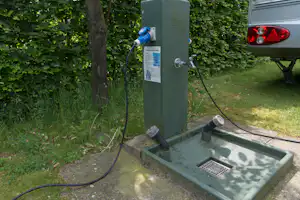 Wasseranschluss am Campingplatz mit Zapfhahn und Schlauch, daneben ein Spülbecken auf Beton, im Grünen mit Hecke und Rasen.