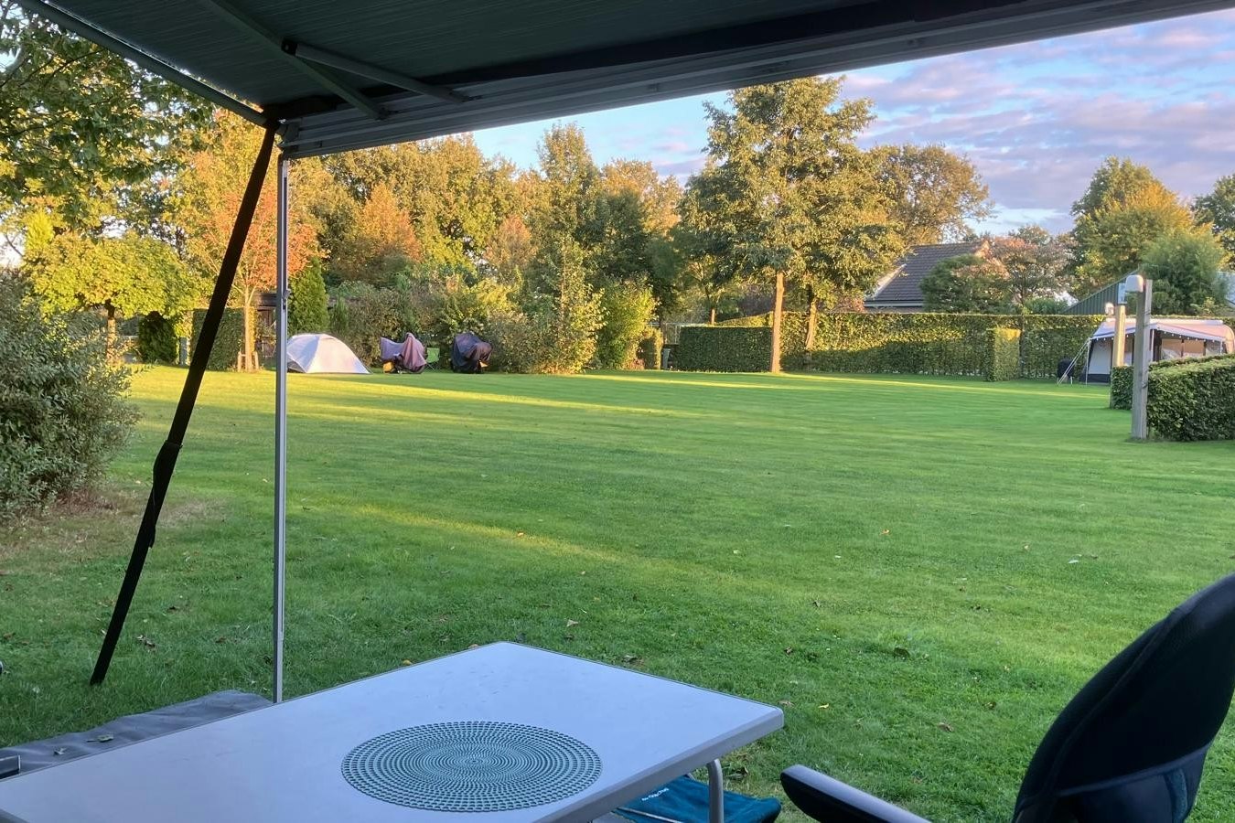 Blick vom Campingplatz unter einem Sonnenschirm auf einen gepflegten Rasen, Sitzgelegenheiten und Zelte im Hintergrund.