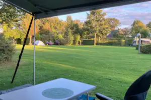 Blick vom Campingplatz unter einem Sonnenschirm auf einen gepflegten Rasen, Sitzgelegenheiten und Zelte im Hintergrund.