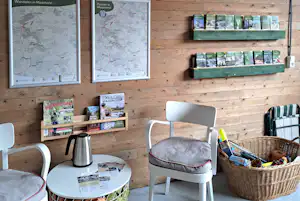 Informationswand mit Karten und Broschüren in einer Holzlounge, daneben Sitzgelegenheiten, Kaffeekanne und Korb auf dem Tisch.