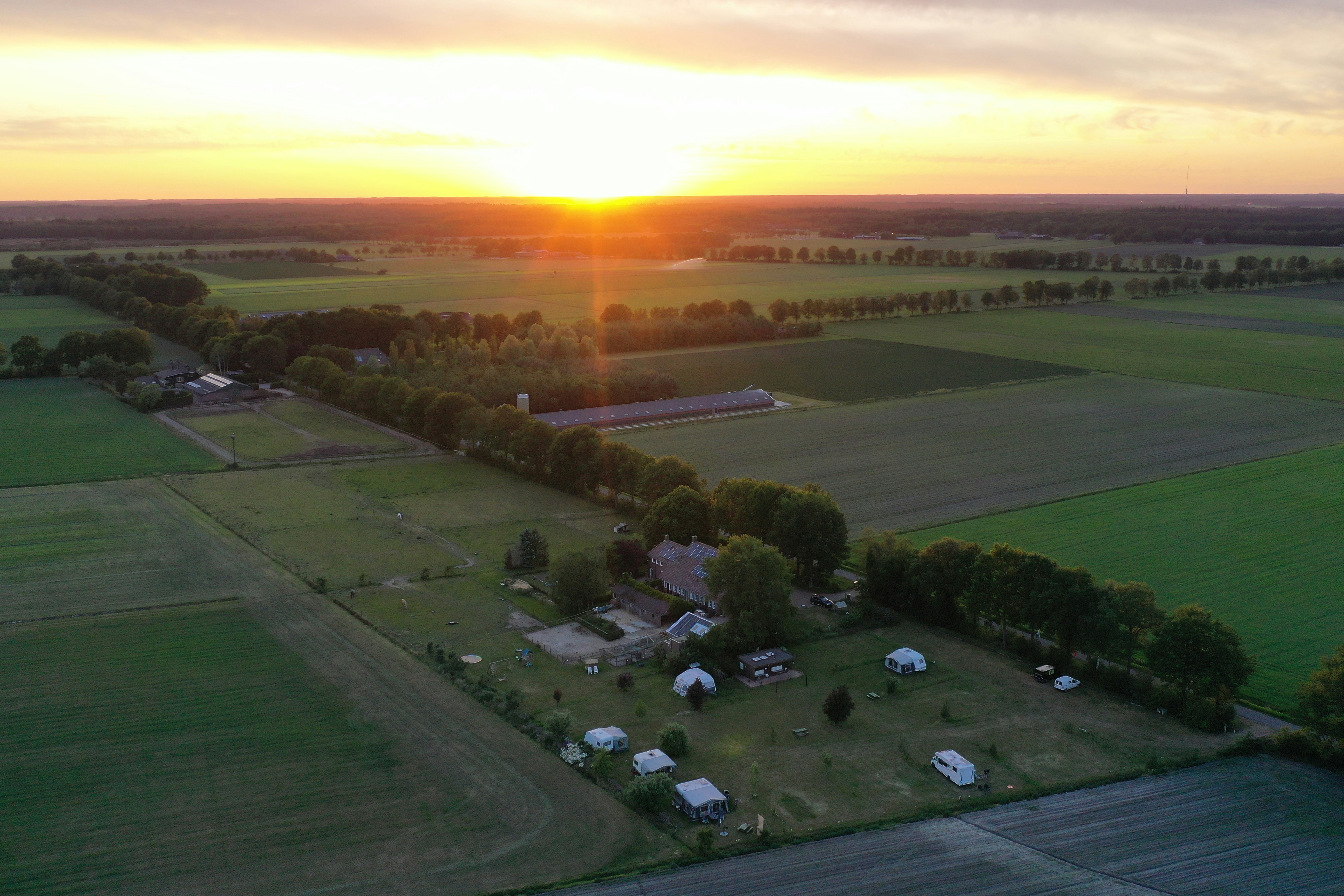 Luftaufnahme eines Campingplatzes mit Stellplätzen und Wohnwagen inmitten von Feldern, Wegen und Bäumen bei Sonnenuntergang.