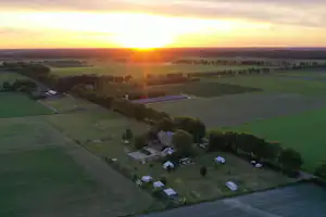 Luftaufnahme eines Campingplatzes mit Stellplätzen und Wohnwagen inmitten von Feldern, Wegen und Bäumen bei Sonnenuntergang.