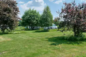 Campingplatz auf der Wiese mit Wohnwagen und weißem Transporter, umgeben von Bäumen und Hecken.