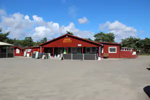 Rotes Holzgebäude mit Rezeption und Schildern, davor Stellplatz mit Absperrungen, im Hintergrund Bäume und blauer Himmel.