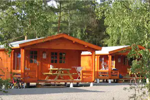 Holzhaus mit Veranda, Sitzbank und Tisch vor dem Eingang, umgeben von Bäumen und Grün im Campingbereich.