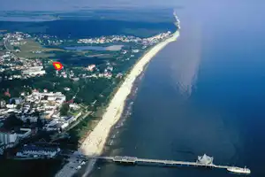 Luftaufnahme der Küste mit langem Sandstrand, Hafen und Promenade, dahinter Häuser und Grünflächen.