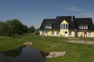 Gelbes Campinghaus mit Terrasse, Holzgeländer und Schaukel vor einem kleinen Teich im Grünen.